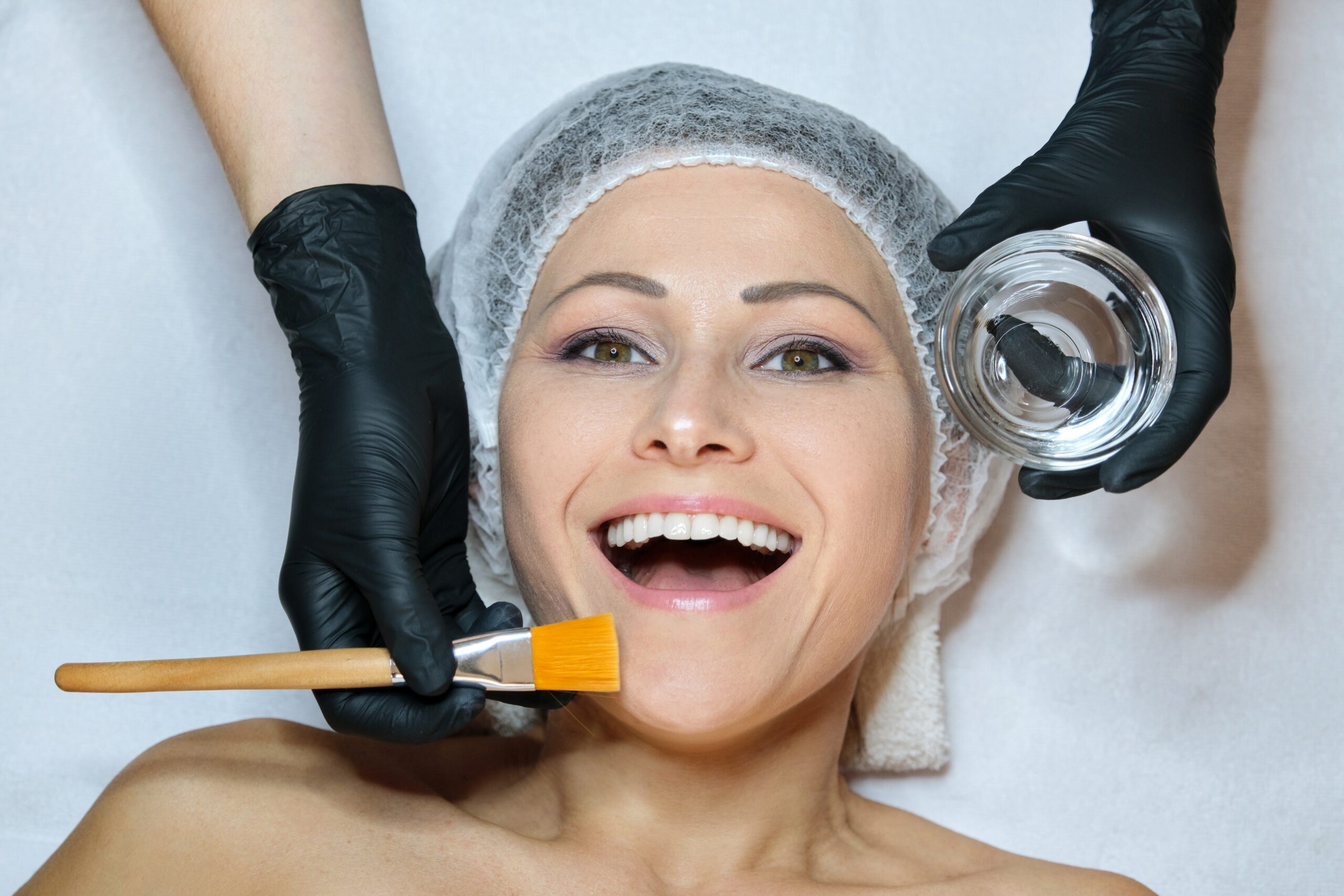 Closeup beautiful smiling happy mature woman lying in beauty salon, hands of cosmetologist woman with brush on patient face, anti-aging cosmetics and procedures